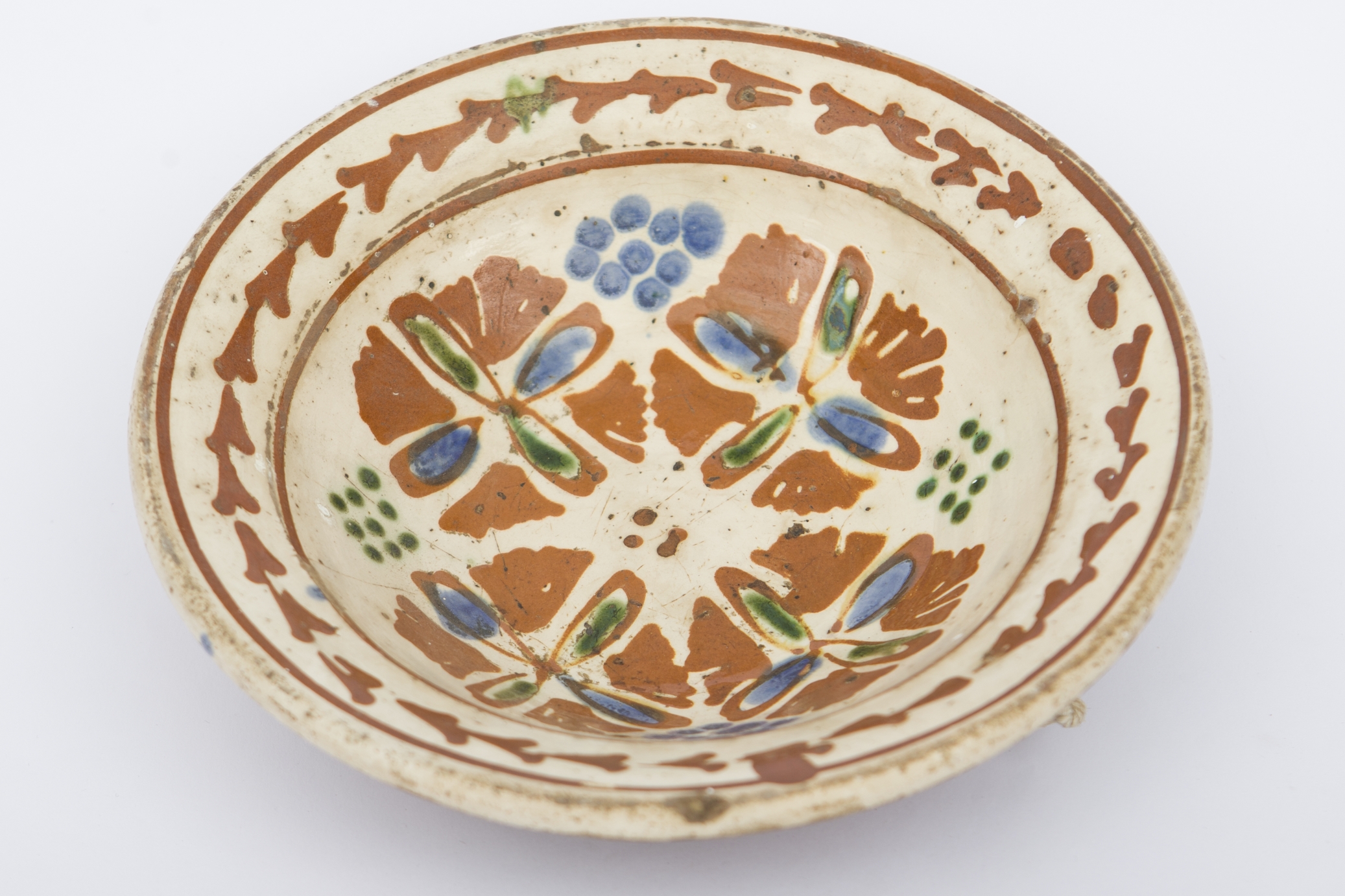 Piesă ceramică smălțuită la interior, de dimenisuni reduse, având formă tronconică. Peste stratul crem de angobă a fost pictat decorul geometrizant și fitomorf stilizat. Pe fundul vasului au fost realizate patru flori cu petale maro, mijlocul verde și albastru, fiind mărginite de patru grupuri a câte nouă puncte verzi și albastre (două câte două, în diagonală). Marginal, pe buza piesei au fost pictate, între două linii circulare drepte un șir de „V-uri” succesive. Farfuria era folosită în gospodărie pentru decorarea interioarelor sau servirea hranei. Prezintă uzură funcțională: Cromatică: fond: cărămiziu; crem; motive: maro; verde; albastru.