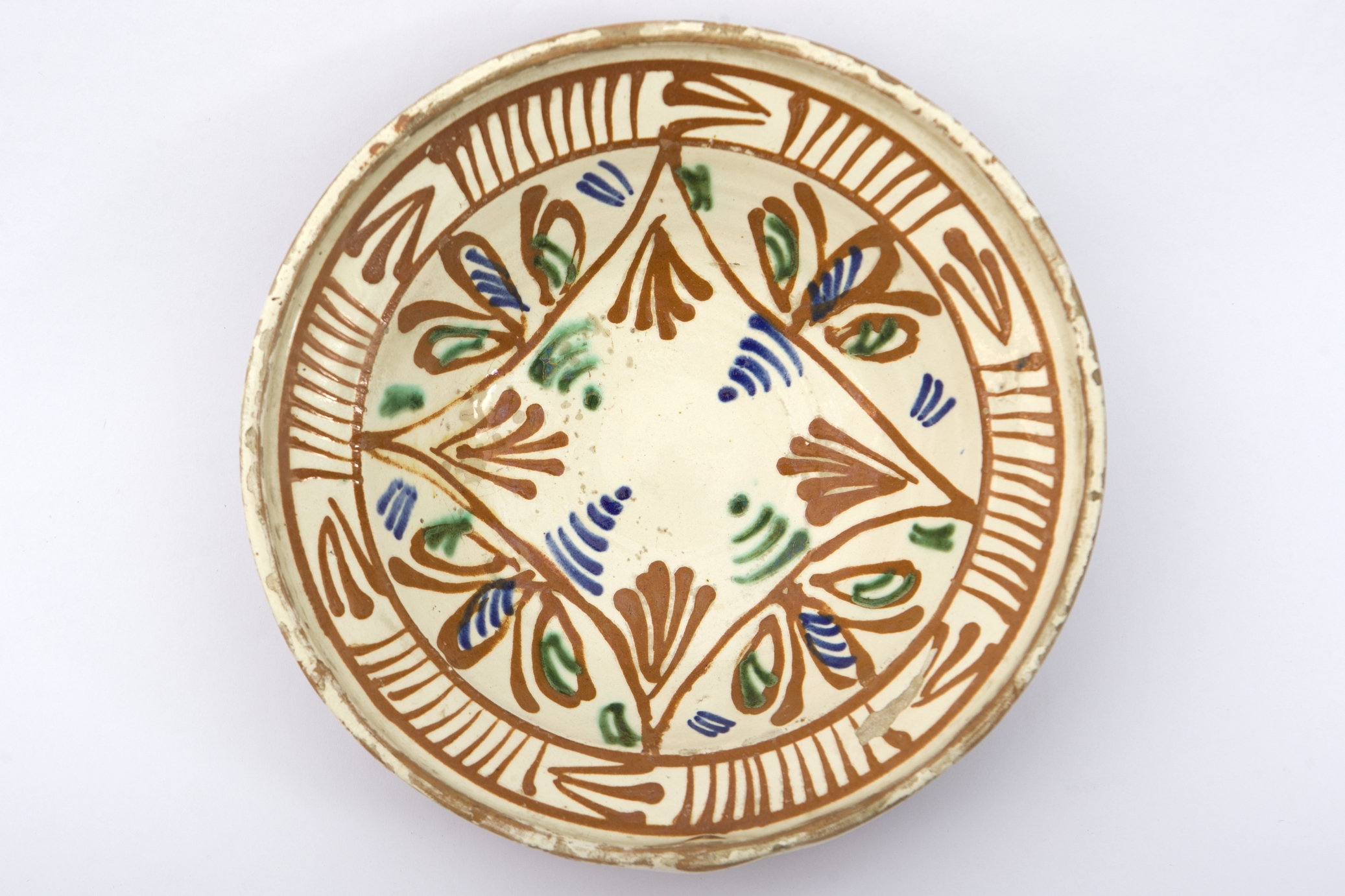 Piesă ceramică smălțuită la interior, de formă tronconică, cu baza plată, pereții evazați și buza profilată. Decorul fitomorf și geometrizant stilizat, dispus pe toată suprafața interioră a farfuriei, a fost pictat peste un strat gros de angobă, de culoare crem. Pe fundul vasului au fost pictate: o formă patrulateră, cu colțurile pronunțate, în interiorul căruia au fost realizate frunze și linii semicirculare. În exteriorul patrulaterului alte patru flori, au fost încadrate între cele patru colțuri. Marginal, pe buza piesei au fost pictate șase grupuri, din linii verticale, despărțite de alte șase grupuri de linii orizontale și oblice, unite la unul din capete. Farfuria era folosită în gospodărie pentru decorarea interioarelor sau pentru servirea hranei. Prezintă uzură funcțională. Cromatică: fond: cărămiziu (exterior); alb (interior); motive: roșu; albastru; verde.