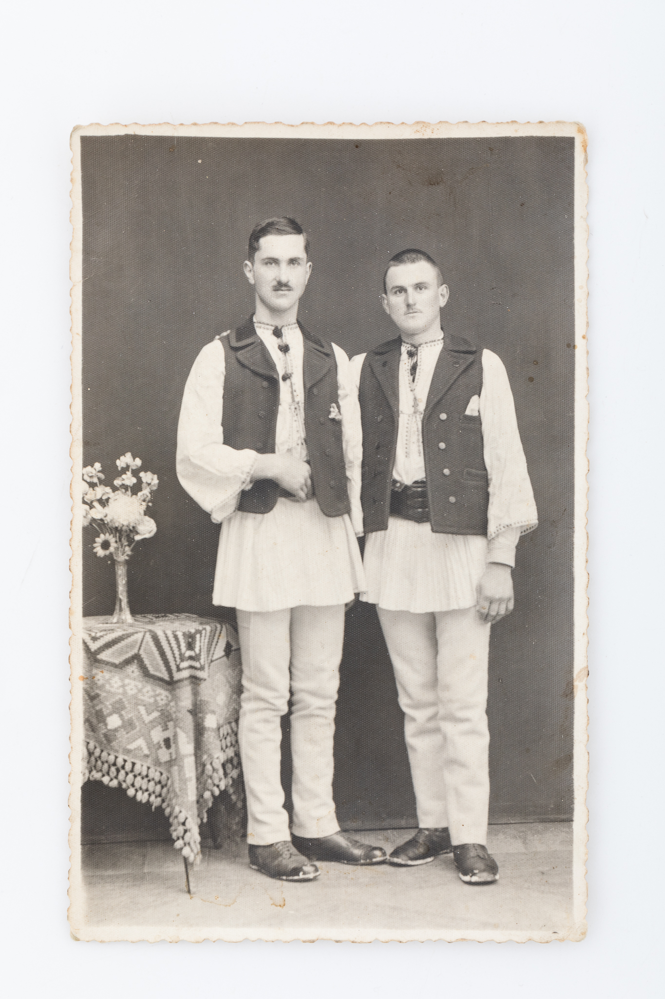 Fotografie de atelier, cu elemente de recuzită: masă, vază cu flori, față de masă. Imaginea este imprimată pe hârtie fotografică, cu gelatino-bromură de argint, în format Carte poștală, D.O.P.. Marginile hârtiei sunt zimțate. V: Rubricatură de carte poștală. Text imprimat, în limba franceză: „Carte postale”. Însemnare manuscrisă: „Comșa / Alisandru Hanzu / Hanzu Ilie”. Număr de inventar MCPT 2697.